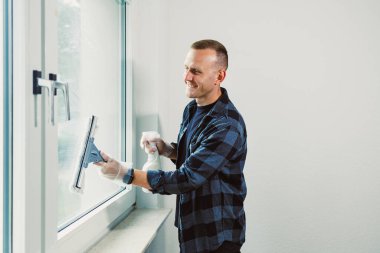 Neşeli genç bir adam doğal ışıkla dolu modern bir odada büyük bir pencereyi temizliyor. Bir bez ve bez kullanıyor, eldiven giyerken görevinden zevk alıyor..