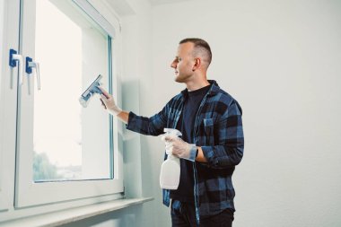 Eldiven giyen bir adam büyük bir pencereyi cam sileceği kullanarak temizlemeye odaklanır. Güneş ışığı odaya akar ve evde bakımını yaparken iç güzelliğin aydınlanmasını sağlar..