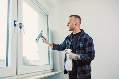 Eldiven giyen bir adam elinde sprey şişeyle pencereyi temizler. Güneş ışığı odayı aydınlatır ve temiz atmosferi güçlendirir..