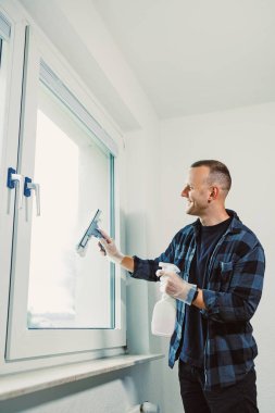 Neşeli bir adam cam silici ile temizliyor, eldiven ve ekose gömlek giyiyor. Işıkla dolu odada taze beyaz duvarlar var. Hoş bir atmosfer yaratıyor..