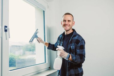 Neşeli bir adam pencereyi temizlemek için cam sileceği kullanıyor, diğer elinde de sprey şişesi var. Güneş ışığı odayı dolduruyor, çabalarını vurguluyor ve mekanı aydınlatıyor..