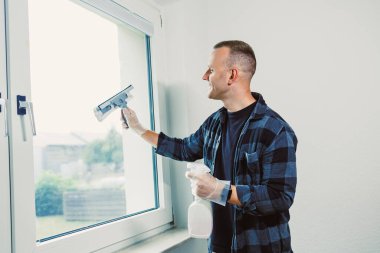 Doğal ışıkla dolu neşeli bir odada, bir adam, temiz bir atmosfer yaratmak için, bir sprey şişesi ve cam sileceği kullanarak, mutlu bir şekilde pencereyi temizliyor..
