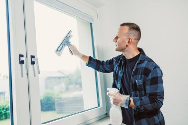 Temiz, aydınlık bir odada elinde sprey şişeyle pencereyi temizleyen bir adam. Güneş ışıl ışıl parlıyor, uzayın neşeli atmosferini arttırıyor..