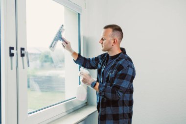 Bir adam parlak bir odada dikilirken pencereyi titizlikle temizler. Güneş ışığı içinden akar ve netliği sağlamak için gayretle çalışırken odaklanmış yüz ifadesini aydınlatır..