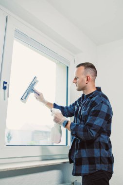 Eldiven giyen bir adam, doğal ışıkla dolu çağdaş bir odada pencereyi özenle temizler, temizlik rutinini gösterir ve düzenli bir ev bakımına odaklanır..