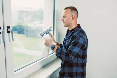 Ekose gömlekli bir adam odaklanma ve özenle büyük bir pencereyi temizler. Odaya giren parlak ışığı artırmak için temizleme solüsyonu sıkar ve yeni bir atmosfer yaratır..