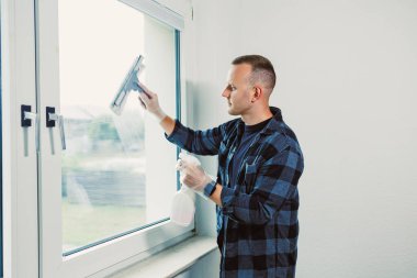 Bir adam parlak bir odada dikilirken pencereyi titizlikle temizler. Güneş ışığı içinden akar ve netliği sağlamak için gayretle çalışırken odaklanmış yüz ifadesini aydınlatır..