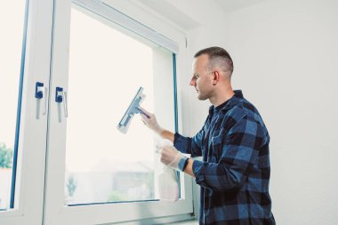 Mavi kareli gömlek giyen bir adam pencereyi titizlikle temizliyor, iç mekânda akan parlak ışığın keyfini çıkarırken iç kısımları silmek için bir cam sileceği kullanıyor..