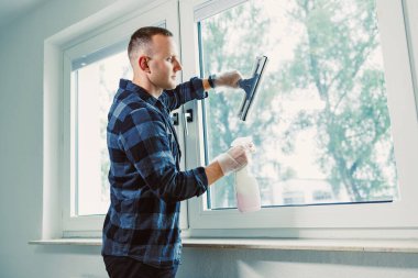 Damalı gömlekli bir adam parlak bir odada pencereleri silmek için cam sileceği kullanır. Doğal ışık ile dolu çağdaş bir ev ortamında sakin bir atmosfer yaratır..