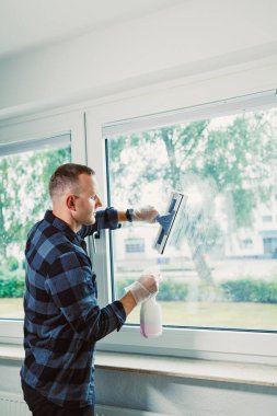 Ekose gömlekli bir adam pencereyi titizlikle temizler, dışarıda temiz bir görüntü sağlar. Güneş ışığı odayı dolduruyor, odaklanmış ifadesini vurguluyor..