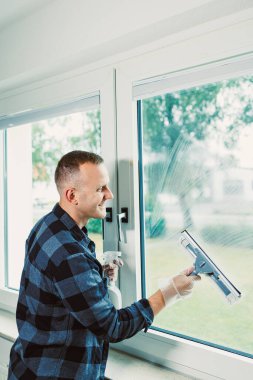 Kareli gömlek giymiş bir adam pencereyi silecekle temizlerken gülümsüyor, odayı suyla dolduran güneş ışığının tadını çıkarıyor ve içeri taze bir esinti giriyor..