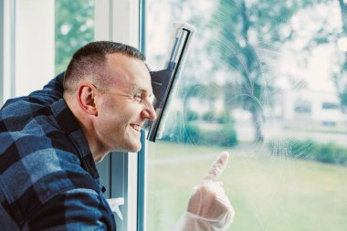 Bir insan pencereyi temizlerken gülümser, dışarıdaki taze manzaranın tadını çıkarır. Güneş ışığı sahneyi aydınlatır, neşeli ve üretken bir atmosfer yaratır..