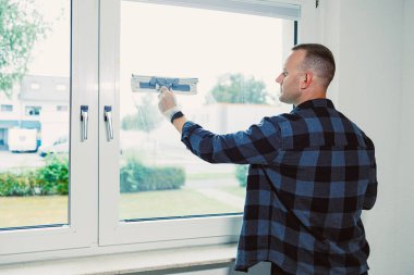 Kareli gömlekli bir adam iyi ışıklandırılmış bir odada pencereyi dikkatlice temizler, dışarıdaki yemyeşil bahçeyi ortaya çıkarır. Konsantrasyonu düzenli bir evin önemini vurguluyor..