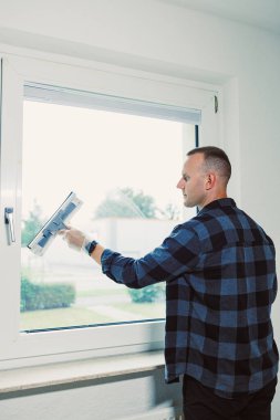 Kareli gömlekli bir adam büyük bir pencereyi silmek için pencere temizleme aleti kullanır. Güneş ışığı parlak boşluğu doldurur, temiz camı ve yemyeşil bahçeyi aydınlatır..