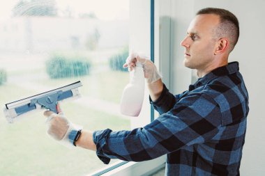 Bir adam kareli gömlek ve eldiven giyerken bir cam sileceği ve sprey şişe kullanarak büyük bir pencereyi dikkatlice temizler. Güneş ışığı aydınlık odayı dolduruyor, temizliği arttırıyor..