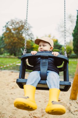 Parkta neşeli bir şekilde sallanan genç bir çocuk, parlak sarı yağmur botları ve güzel sonbahar ağaçlarıyla çevrili bir şapkayla oyun ve doğa ruhunu kucaklıyor..