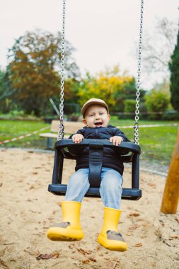 Genç bir çocuk, parlak sarı çizmeler ve rahat bir ceket giyerek parkta sallanmaktan hoşlanır. Hava kıtır kıtır, renkli yapraklar etrafa saçılmış, neşeli bir atmosfer yaratıyor..