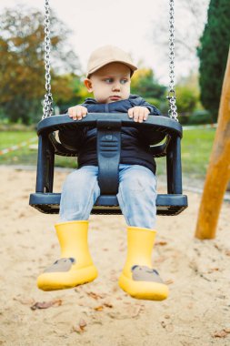 Küçük çocuk parkta sallanıyor, şapkası ve sarı çizmeleri var. Sahne, keyifli bir masumiyeti ve dışarıda oynamanın güzelliğini hoş bir günde yakalar..