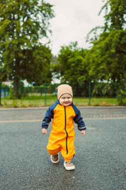 Göz alıcı sarı yağmur kıyafetleri giymiş canlı bir çocuk yemyeşil ve bulutlu gökyüzü ile çevrili bir parkta kaldırımlı bir yolda neşe içinde yürüyor, saf mutluluğu somutlaştırıyor..