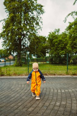Göz kamaştırıcı turuncu tulum giymiş genç bir çocuk canlı ağaçlarla dolu bir parkta kıvrımlı bir patika boyunca yürüyor. Eğlenceli an, çocukluk keşiflerinin özünü yakalar..