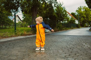 Parlak turuncu yağmurluk giymiş neşeli bir çocuk kaldırımlı bir sokakta neşeli bir şekilde dönüyor. Bulutlu gökyüzü, yeşil ağaçlar ve evlerle çevrili o ana büyülü bir his katıyor..