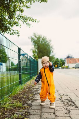 Canlı turuncu tulum giymiş küçük bir çocuk kaldırımlı bir yolda güvenle yürüyor. Meraklı çocuk ileriyi işaret ediyor, serin ve bulutlu günün tadını çıkarıyor. Yeşillik bölgeyi sarmış durumda..