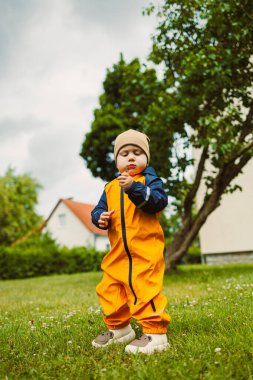 Parlak turuncu bir elbise giymiş neşeli bir çocuk, bulutlar tepemizde sürüklenirken, toplaması için çiçek arayan bir bahçe macerasının tadını çıkarıyor. Yeşil çimenler küçük beyaz çiçeklerle benekli..
