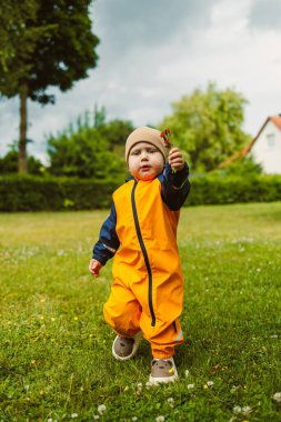 Neşeli genç bir çocuk parlak turuncu bir tulumla çimenli bir parkta yürüyor, canlı bir çiçek tutuyor. Güneş bulutların arasından bakıyor, canlı bir atmosfer yaratıyor..
