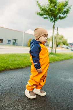 Parlak turuncu tulum giymiş meraklı bir çocuk kaldırımda duruyor. Serin bir meltem, bulutlu bir günde etraflarını gözlemlerken berelerini hafifçe hareket ettirir..