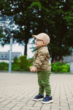 Neşeli bir çocuk yeşil ağaçlarla çevrili bir parkta duruyor, kahverengi bir şapka ve açık renk bir ceket giyiyor. Çocuk heyecanlı ve dışarıyı keşfetmeye hazır görünüyor..