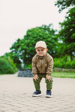 Kahverengi ceketli ve şapkalı küçük bir çocuk ağaçlarla çevrili yemyeşil bir parktaki kaldırımlı bir yolda neşeli bir şekilde çömelmiş, gençlik enerjisi ve merakını gösteriyor..
