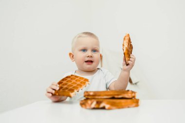 Neşeli bir çocuk heyecanla gülümseyen iki parça altın waffle 'ı havada tutuyor. Masanın üzerinde bir yığın taze waffle oturuyor, parlak bir mutfakta neşeli bir sahne yaratıyor..