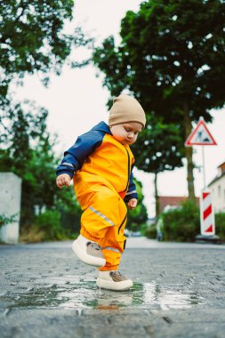 Parlak sarı yağmurluk giymiş küçük bir çocuk su birikintisine atlayıp etrafa su sıçratıyor. Ağaçlar ve sessiz bir sokak Yağmurlu bir öğleden sonra neşeli bir sahne hazırladı.