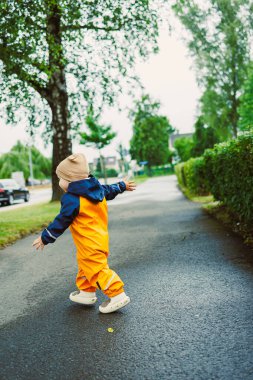 Turuncu ve mavi yağmurluklu küçük bir çocuk ıslak bir yolda neşeyle yürüyor, kolları açık, yemyeşil ve gökten yağan yumuşak yağmurlarla çevrili..