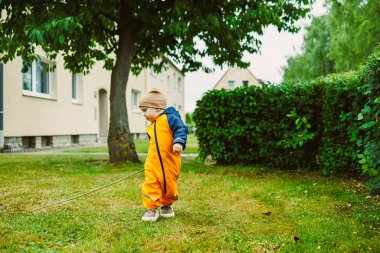 Canlı turuncu takım elbiseli bir çocuk yeşil bir bahçede neşeli bir şekilde dolaşır, doğayı keşfeder ve çimenli araziler ve yakındaki çalıların arasında bir sopayla oynar..