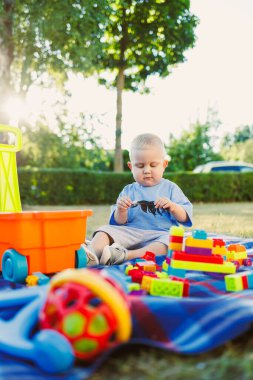 Parktaki mavi bir battaniyenin üzerinde oturan genç bir çocuk, parlak blokları olan bir binaya odaklanmış. Yakınlarda, bir oyuncak arabası ve renkli bir top onun açık gökyüzünün altındaki oyun saatine neşe katıyor..