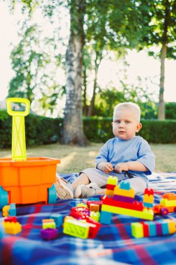 Parkta güneşli bir günde, meraklı bir bebek kareli bir battaniyeye oturur. Renkli yapı taşları etrafa saçılmış durumda, ve yakınlarda parlak turuncu bir araba duruyor, manzaraya neşe katıyor..