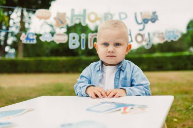 Neşeli genç bir çocuk masaya oturur, düşünceli bir şekilde kameraya bakar, mavi süslemelerin arasında, arka planda yeşillik, neşeli atmosferi yükseltir..