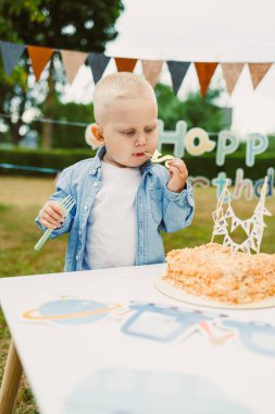 Açık renk saçlı bir çocuk neşeli bir doğum günü kutlaması sırasında bir dilim pastanın tadını çıkarıyor. Renkli afişler ve dekorasyonlar parkta şenlikli bir atmosfer yaratıyor..