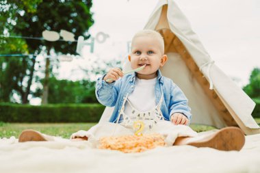 Neşeli bir çocuk, dekoratif bir çadırın yanındaki battaniyeye oturmuş, bir kaşıkla bir dilim pastanın tadını çıkarıyor, yeşil ve şenlikli bir doğum günü kutlaması için dekoratif süslerle çevrili..