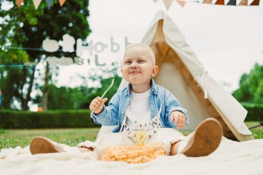 Neşeli bir bebek sıcak bir battaniyeye oturup canlı bir parkta renkli bir pastanın tadını çıkarıyor. Neşeli kiraz kuşu hafif esintide hışırdıyor, neşeli havayı artırıyor..