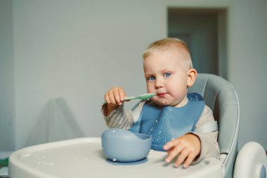 Parlak mavi gözlü genç bir çocuk yüksek bir sandalyeye oturur, mutlu bir şekilde beslenir ve yemeklerle kirlenir. Güneş ışığı basit, temiz mutfağı doldurur, neşeli anı ön plana çıkarır..