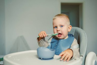 Neşeli genç bir çocuk yüksek bir sandalyede oturur, kaşıkla yemek yer ve temiz ve modern bir mutfak ortamı ile çevrili parlak mavi gözleri sergiler..