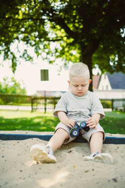 Genç bir çocuk kumlu bir alanda oturuyor, mavi oyuncak kamyonuna odaklanmış. Güneş ışığı ağaçların arasından süzülüyor, sıcak bir öğleden sonra parkta neşeli bir atmosfer yaratıyor, eğlence ve hayal gücüne davet ediyor..