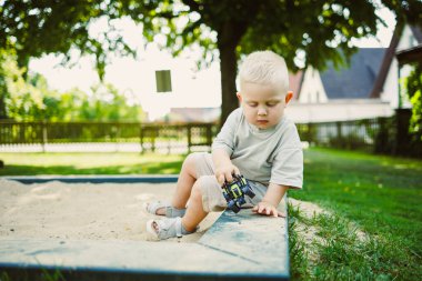 Küçük bir çocuk, bir oyuncak kamyona manevra yaptırmaya odaklanırken bir kum havuzunda oturuyor. Güneş ışıl ışıl parlıyor, neşeli ve huzurlu bir ortam yaratıyor..