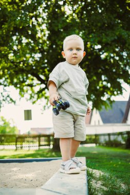 Neşeli bir çocuk oyun parkında duruyor, oyuncak bir arabayı kavrıyor, yemyeşil ağaçlar ve açık mavi gökyüzü ile çevrili, öğleden sonra güneşinin tadını çıkarıyor..