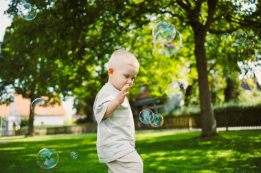 Açık gri bir elbise giymiş genç bir çocuk, neşe ve neşe dolu sıcak, yemyeşil bir parkta renkli baloncukları kovalamaktan ve patlatmaktan hoşlanıyor..