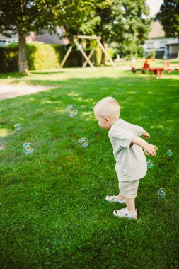 Tıraş olmuş kafası olan genç bir çocuk neşeli bir şekilde ağaçlarla ve oyun alanlarıyla dolu yemyeşil bir parkta baloncukları kovalıyor, güneşli bir günde tasasız bir çocukluğun özünü yakalıyor..