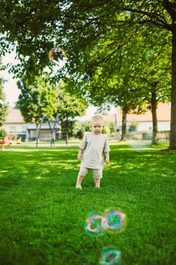 Genç bir çocuk canlı bir parkta çimlerin üzerinde duruyor, sıcak güneş ışığının tadını çıkarıyor ve renkli kabarcıkların havada sürüklenişini seyrediyor. Oyun parkı yakınlarda görünüyor, eğlenceli sahnelere ekleniyor.