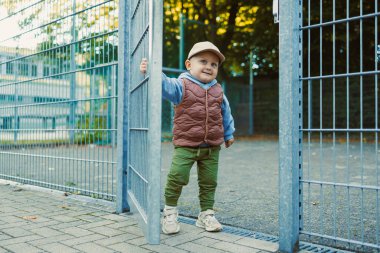 Neşeli genç bir çocuk metal bir kapının yanında duruyor, büyük bir gülümsemeyle onu gururla tutuyor. Samimi bir yelek ve şapka giyiyor, oyun alanının zevklerini keşfetmeye hazır..